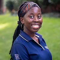 Mark Strauss, with long braided hair and a patterned headband, smiles confidently in his navy blue polo shirt adorned with a logo, as he stands on the grassy area.