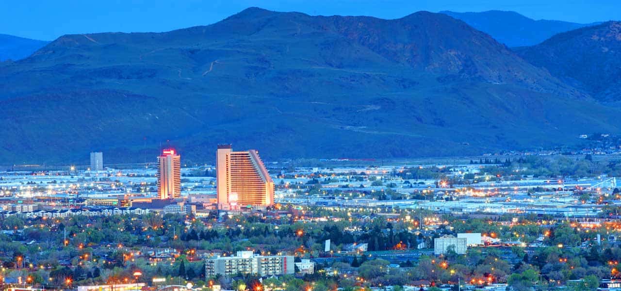 A cityscape at dusk featuring two illuminated high-rise buildings, surrounded by smaller structures and greenery, with mountains in the background—an inspiring setting for the Nevada MPD Annual Meeting.