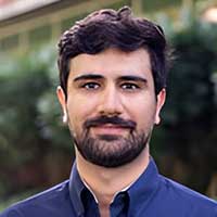 Mark Strauss, a person with short, dark hair and a beard, stands outdoors in a blue shirt. The backdrop of blurred greenery emphasizes his calm demeanor amid nature's embrace.