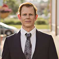 Mark Strauss stands outdoors in an urban setting, clad in a suit and tie. Behind him, a blurred car weaves through the greenery, painting a picture of vibrant city life.