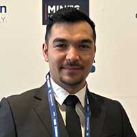 A man in a suit and tie, identified as Mark Strauss, stands confidently in front of a backdrop adorned with logos. He has short dark hair and wears a lanyard.