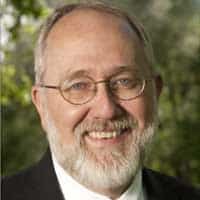 Carl Nesbitt, a middle-aged man with glasses, a gray beard, and a suit, smiles outdoors with greenery in the background.