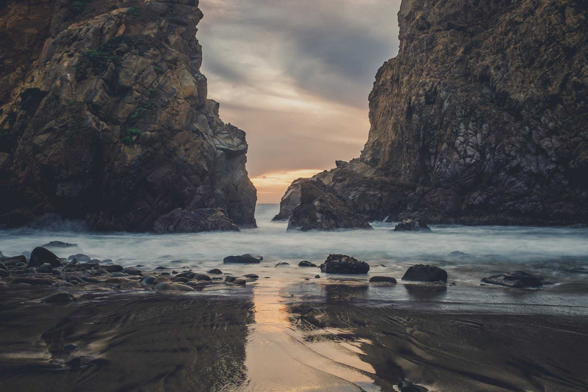 A rocky beach scene at sunset, with large cliffs framing a narrow passage to the sea. Waves gently lap at the shore, reflecting the colors of the sky.