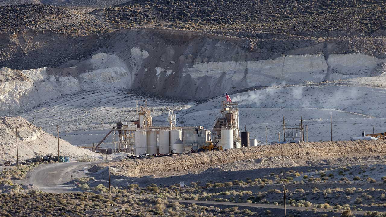 An industrial facility with tall structures and storage tanks is set against a rocky, barren landscape. A road leads to the site, and a flag is visible atop one of the structures.