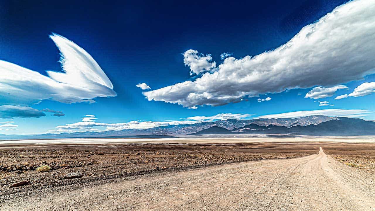 A vast desert landscape with a dirt road, distant mountains, and dramatic cloud formations in a bright blue sky.