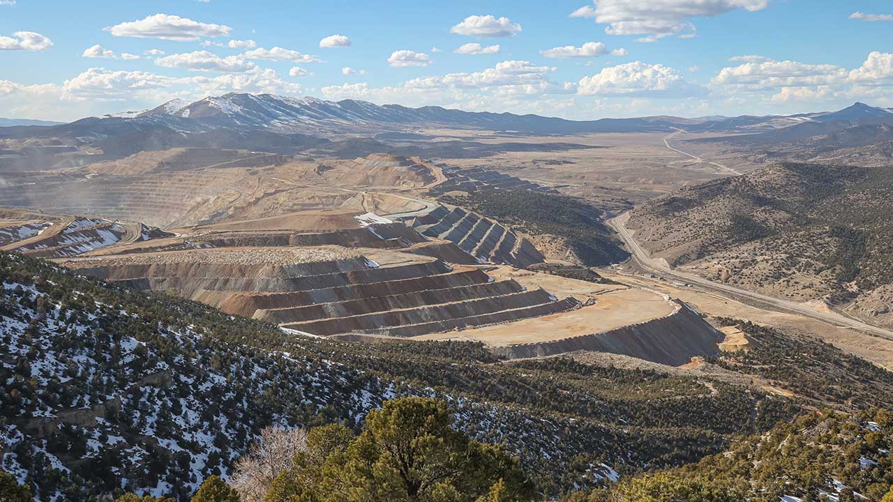 Aerial view of a large open-pit mine with terraced layers, surrounded by rugged mountains and sparse vegetation under a partly cloudy sky.